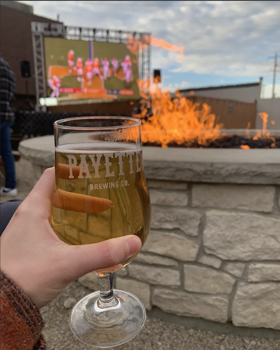 A hand holding a Payette Brewing Co. stemmed pour glass in front of a stone fire pit and a big screen showing a football game