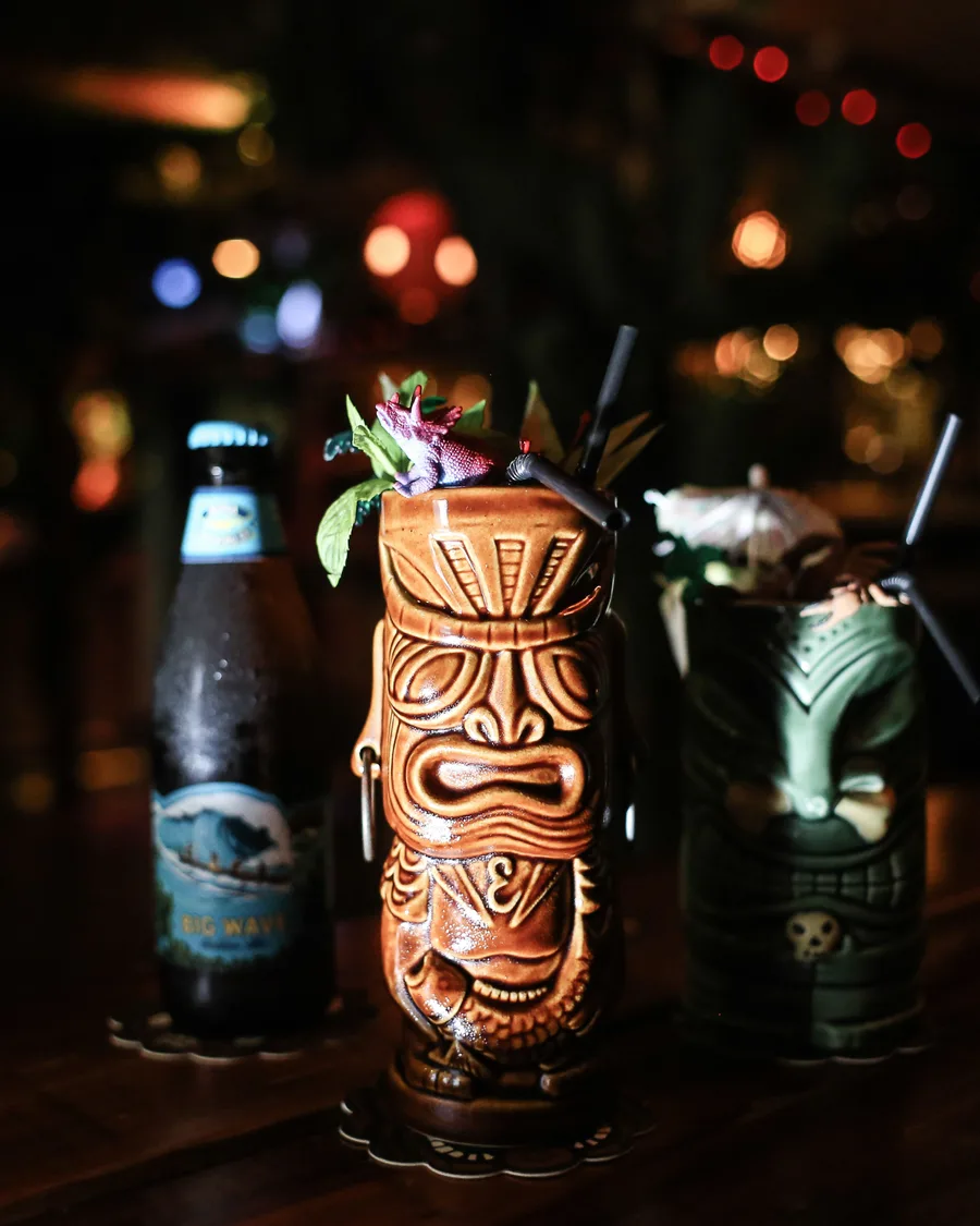 Two carved tiki mugs and a Big Wave beer on a dark bar top, lit by string lights