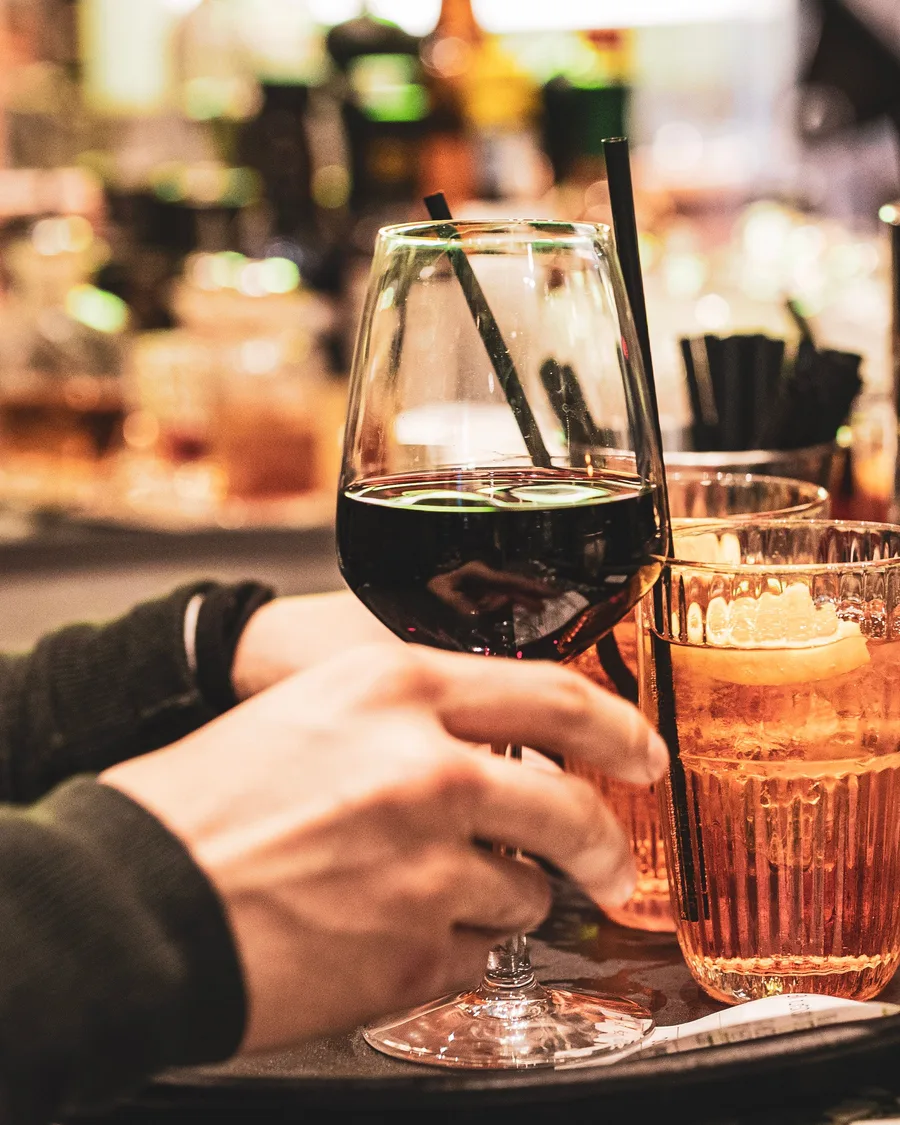 A hand lifting a glass of red wine beside an orange spritz and more drinks on a bar