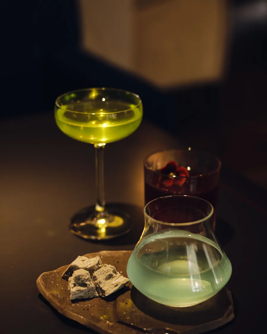 A tasting flight of three cocktails in low light, with a small dessert on a ceramic plate