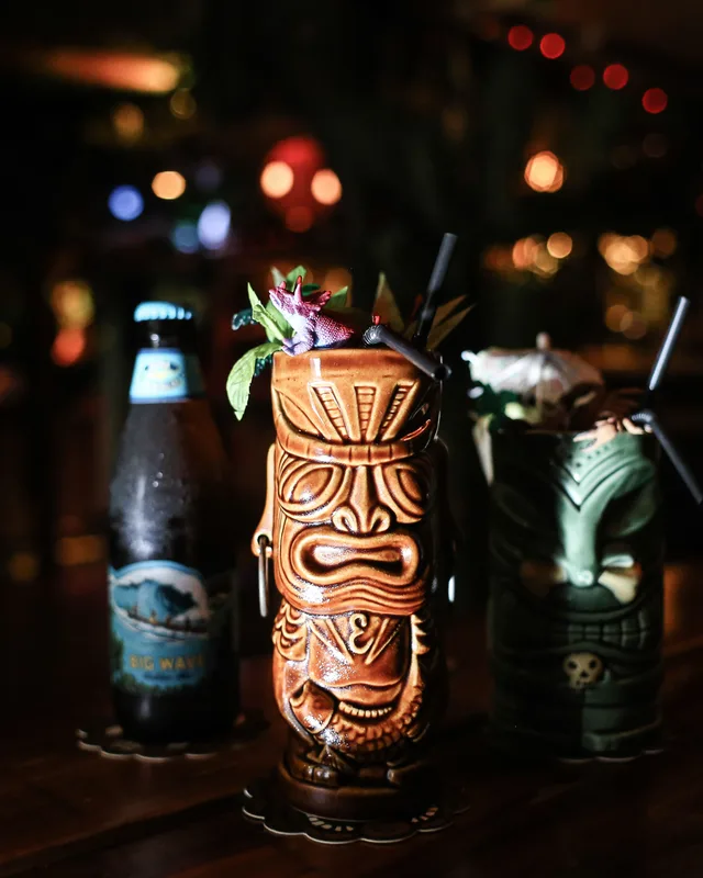 Two carved tiki mugs and a Big Wave beer on a dark bar top, lit by string lights