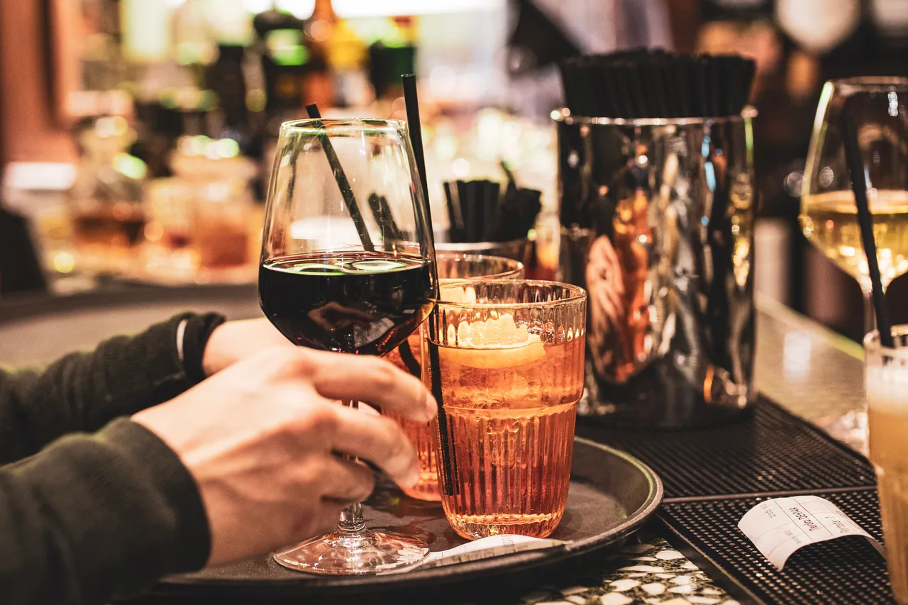 A hand lifting a glass of red wine beside an orange spritz and more drinks on a bar